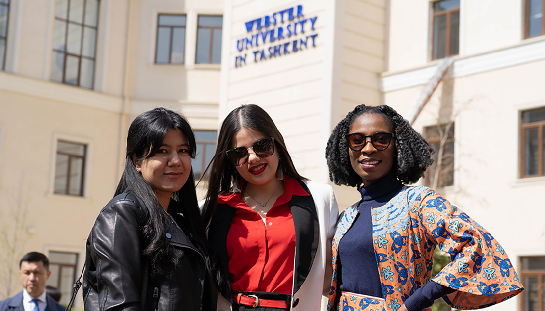 Three women stand together in front of ̳ Tashkent sign with a man standing to the side of them..